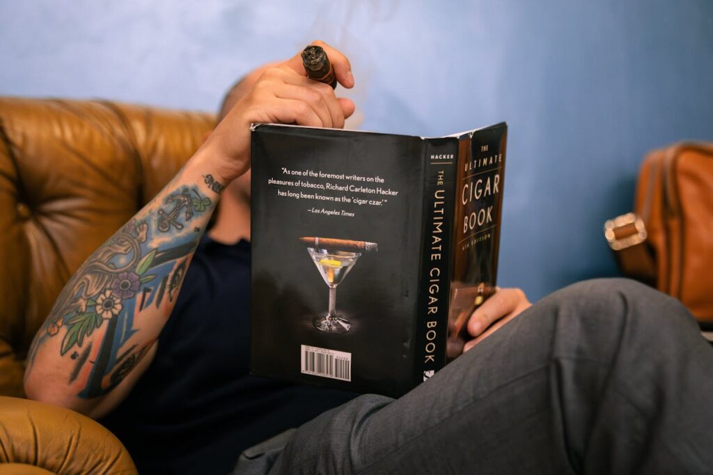 Man reading a book and smoking a cigar on a sofa.