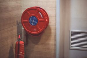 Red fire hose reel and fire extinguisher mounted on a wooden wall indoors.