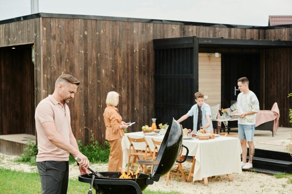 Man grilling near a wooden home while others prepare a meal outdoors.